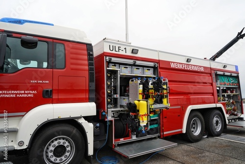 View on red fire-brigade truck with open side door where is visible firefighting equipment. On aft part is written with white letters fire department in German language.