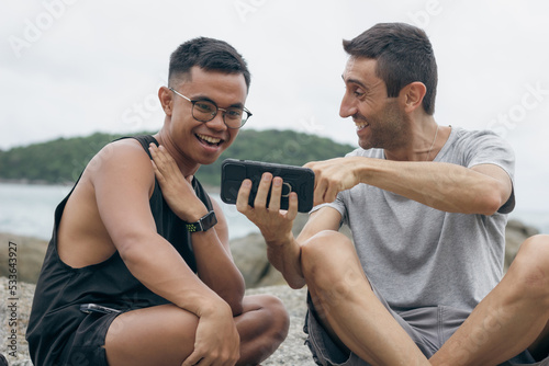 Filipino young man and caucasian guy laughing looking at cellphone outdoors. Couple of male friends enjoying social media on smart phone. Ethnicity diversity, sharing internet joke concepts