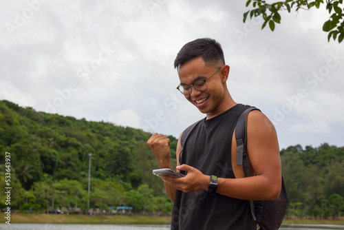 Euphoric Filipino man celebrating sport victory looking at smart phone by lake park. Lucky male person acting exultant holding mobile. On line bet win concept