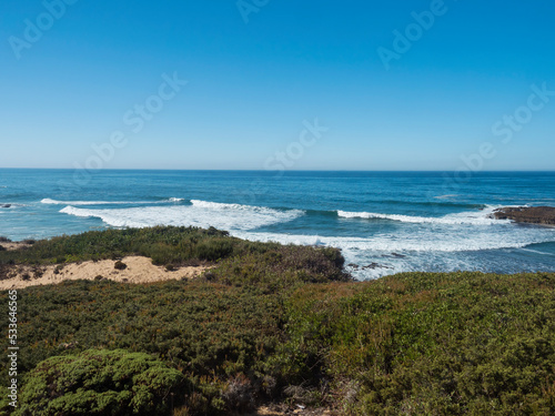 View of wild Rota Vicentina coast, sea shore with ocean waves , sand and green vegetation at sunny day, clear blue sky