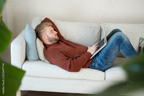 Young caucasian blond curly hair man lying on sofa looking at laptop, using computer,working online at home in modern scandinavian interior.