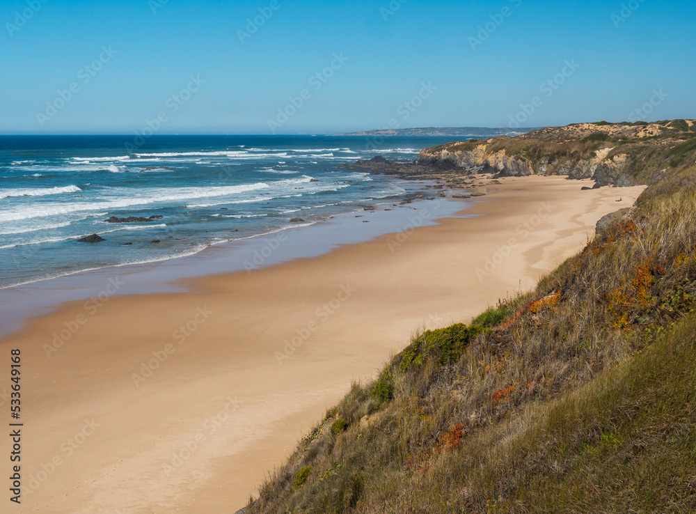 View of empty Praia do Brejo Largo beach with ocean waves, stone, wet golden sand and green vegetation at wild Rota Vicentina coast near Vila Nova de Milfontes, Portugal.