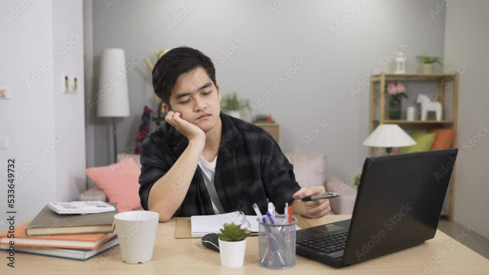 portrait of an absent minded Asian college student is propping face and playing with his pen feeling bored while learning from home in the living room. portrait of an absent minded Asian college student is propping face and playing with his pen feeling bored while learning from home in the living room.