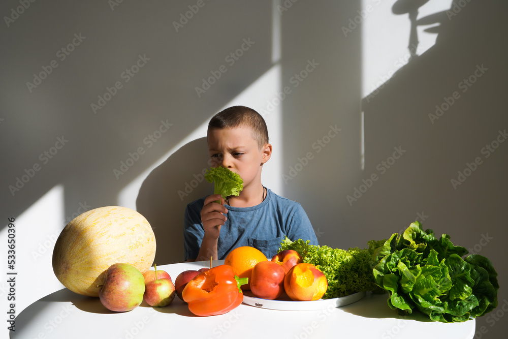 Fototapeta premium Boy sitting at a table with vegetables and fruits in daylight and holding a salad in his hand with a displeased face