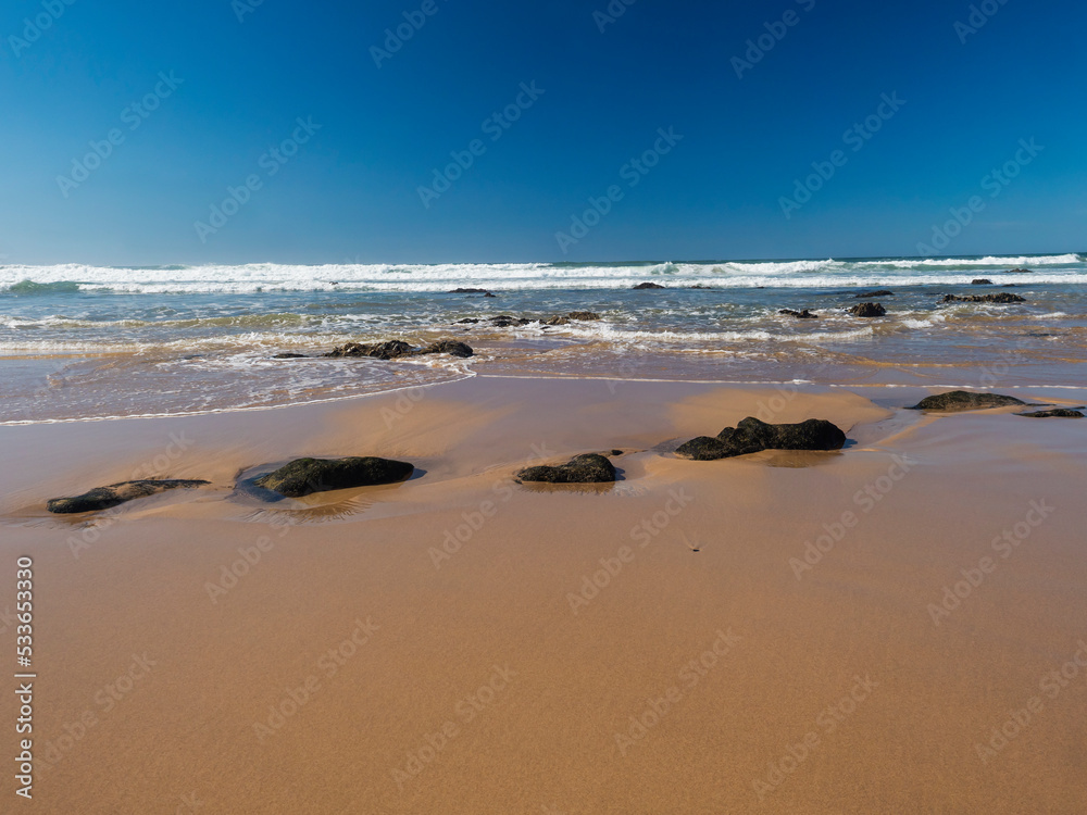 View of empty Praia do Brejo Largo beach with ocean waves and sharp rocks and wet golden sand at wild Rota Vicentina coast near Porto Covo, Portugal.
