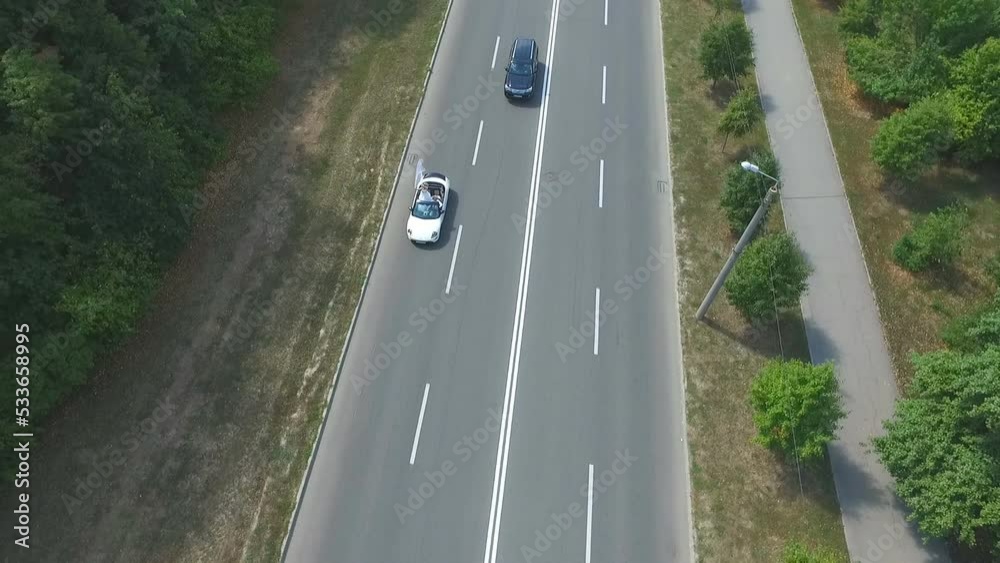 Aerial view of white convertible car riding along country road. Young couple traveling on cabriolet. Happy just married pair enjoying trip together. Flying over auto driving on highway at summer day Aerial view of white convertible car riding along country road. Young couple traveling on cabriolet. Happy just married pair enjoying trip together. Flying over auto driving on highway at summer day