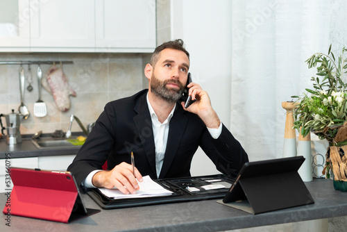 Busy young businessman telephones and takes notes in the kitchen. Handsome business man working at home. Smart working with tablets and sheets during the covid pandemic.