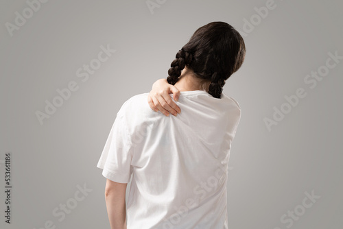 Arthritis and chondrosis. Woman with piggy tails holds her shoulder with her hand, feeling pain. Rear view. Gray background. The concept of injuries of ligaments and joints