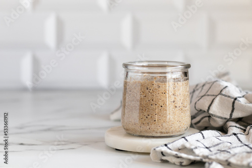 Wheat sourdough in a glass jar on a marble coaster nearly kitchen towel. Ingredient for artisan bread. White background. Copy space photo