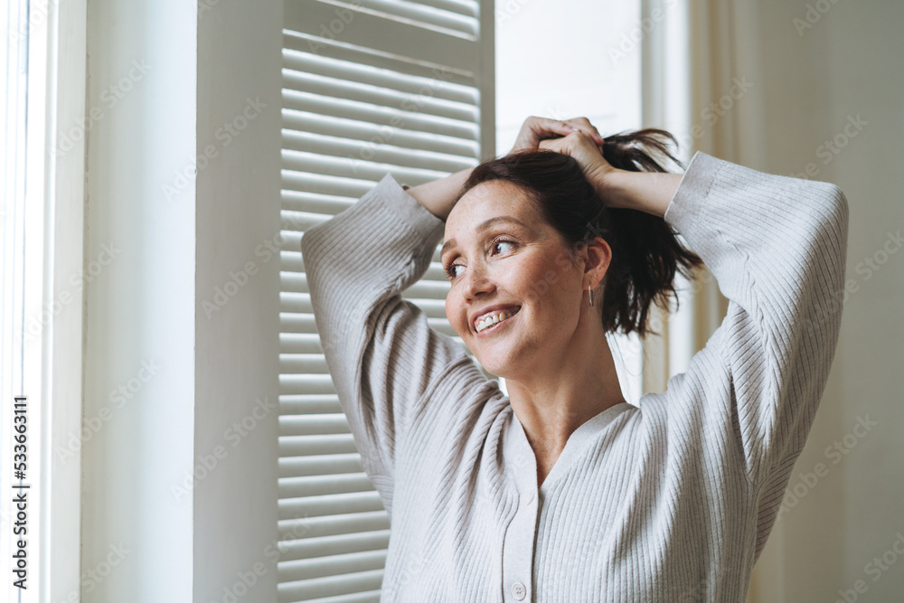 Young beautiful woman forty year with brunette hair in cozy knitted cardigan near window in bright interior at home Young beautiful woman forty year with brunette hair in cozy knitted cardigan near window in bright interior at home