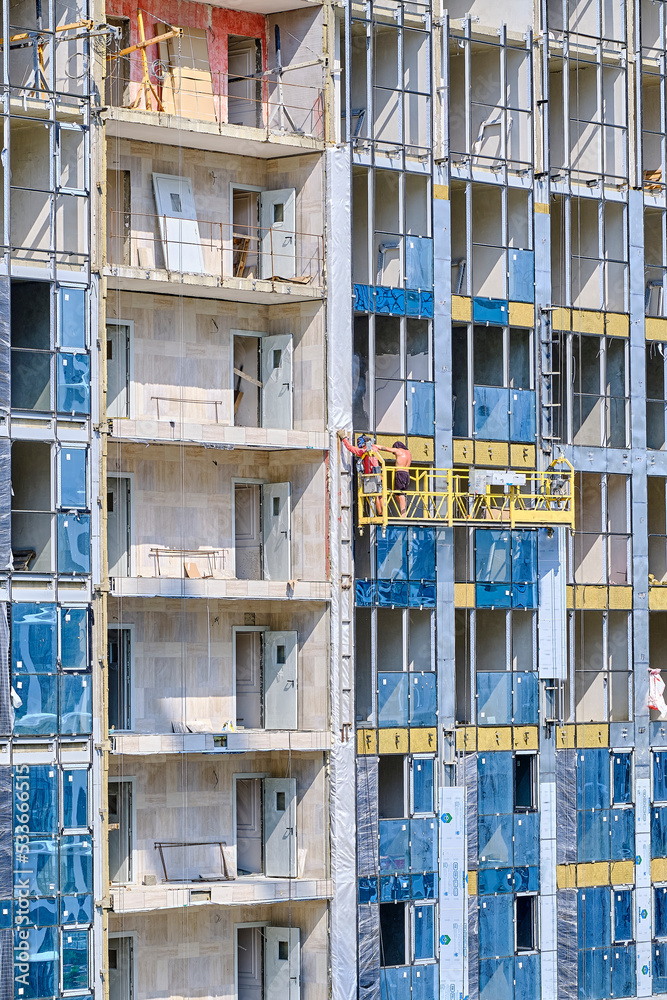 Being on construction cradle, builders insulate walls of facade of multi-storey residential building under construction. Heat saving concept.Vertical footage.