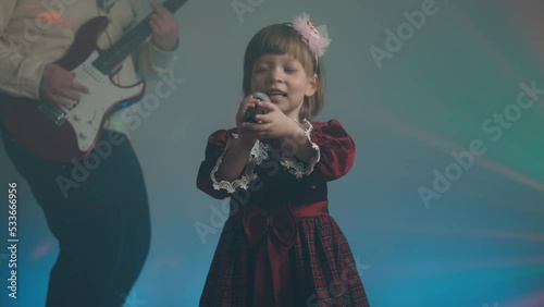Little girl on stage in vintage dress, she sings into microphone and dances, her father plays an electric guitar. Color music is shining and smoke is billowing. Family time, creativity and hobbies