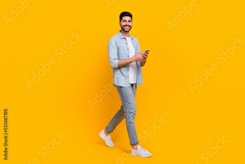 Full body photo of handsome latin young guy model walk hold device read news wear trendy jeans clothes isolated on yellow color background