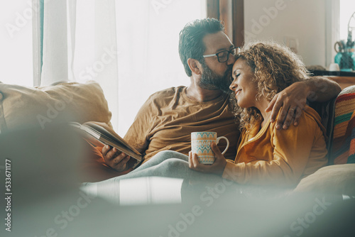 Couple of young adult man and woman enjoy time together sitting on the sofa in indoor leisure activity.. Real life people at home. Male and female relax on the couch reading a book and drinking a tea photo