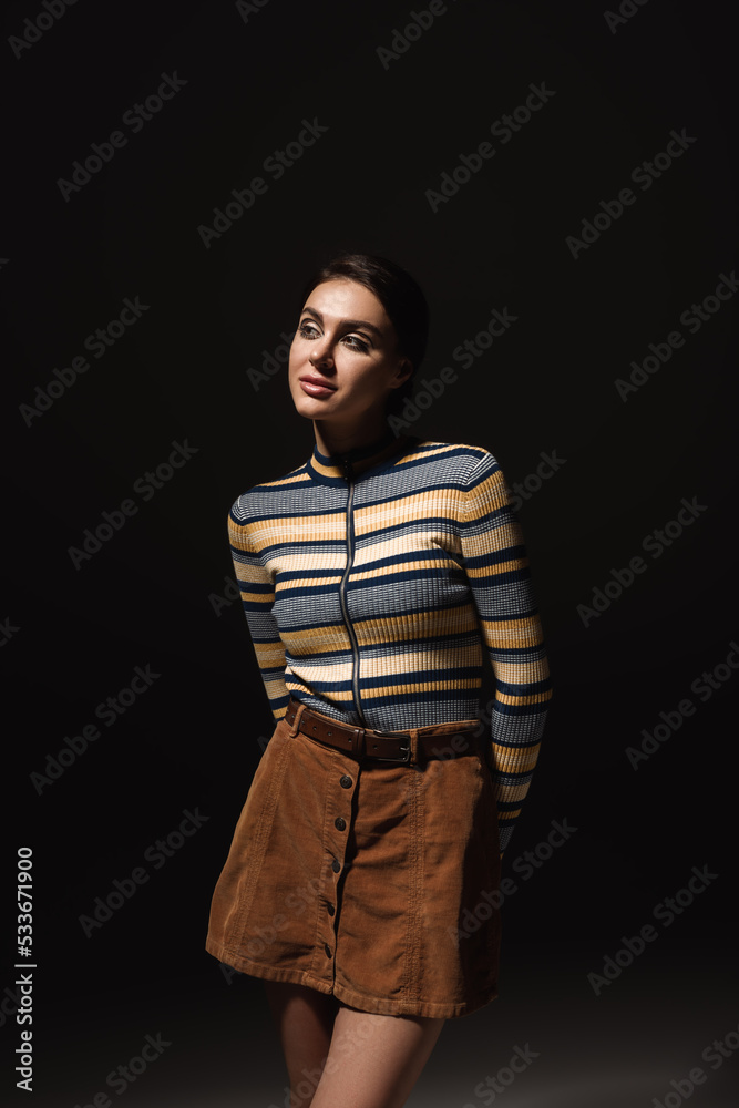 brunette young woman in striped long sleeve turtleneck and skirt looking away isolated on black. brunette young woman in striped long sleeve turtleneck and skirt looking away isolated on black.
