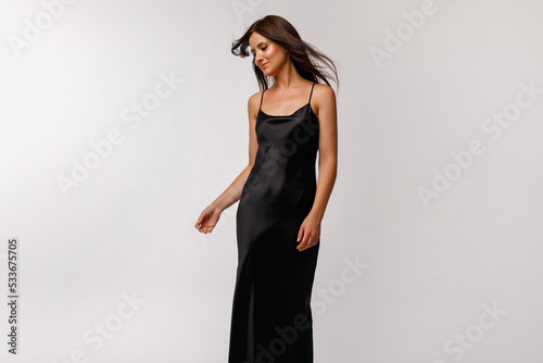 A brunette girl with long hair is standing in a black slip dress on a light background photo