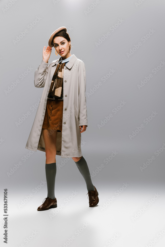 full length of elegant young woman in autumnal trench coat adjusting beret on grey. full length of elegant young woman in autumnal trench coat adjusting beret on grey.