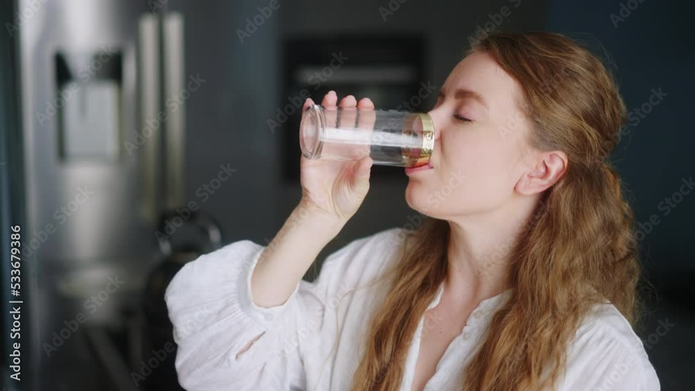 Thirsty smiling young woman standing alone in domestic kitchen drinking water from glass. Girl start new day with healthy life habit, sipping clean mineral natural still water close up view. Thirsty smiling young woman standing alone in domestic kitchen drinking water from glass. Girl start new day with healthy life habit, sipping clean mineral natural still water close up view.