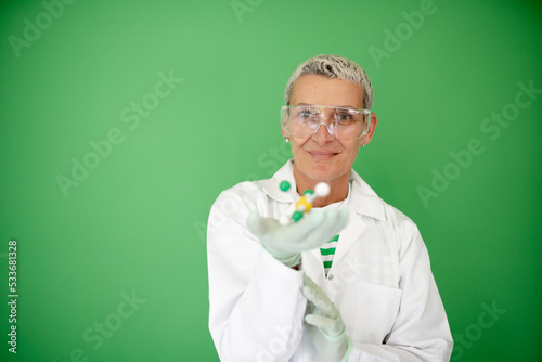female scientist with white coat is holding model of molecule in her hand and is standing in front of green background