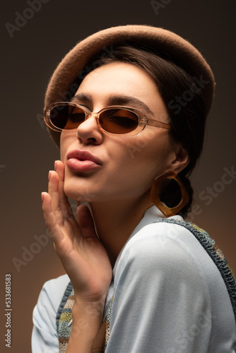 stylish young woman in beret and sunglasses pouting lips on brown.
