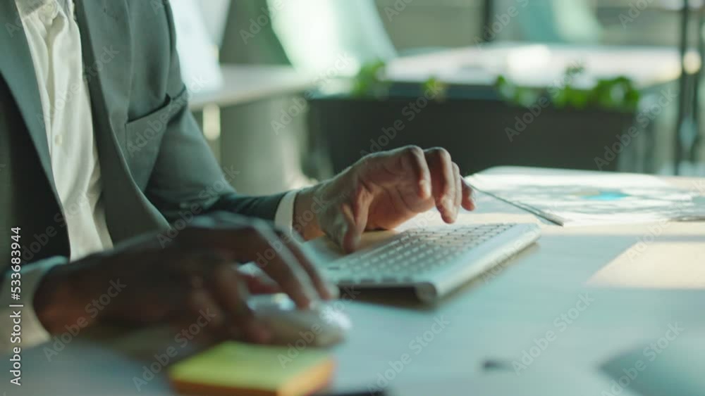 Portrait of good-looking bearded thoughtful middle-aged African man in formal outfit working at computer. Close-up shot of male hands using computer mouse. Indoors