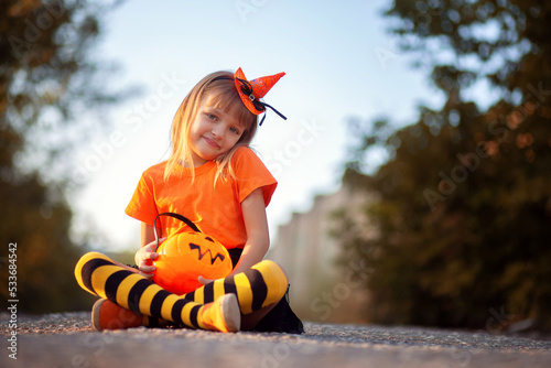 A little girl in a Halloween outfit sits on the ground on the street with a large candy bowl and smiles. Merry Halloween