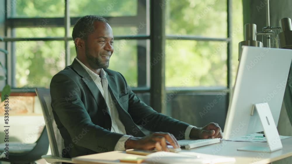 Portrait of thoughtful good-looking African man sitting at workplace with computer and touching face. Shot of smiling successful businessman looking at screen, reading e-mail in office. Daytime