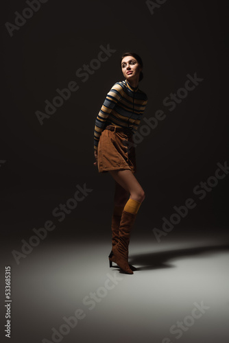 full length of young woman in turtleneck with skirt and boots posing on black.