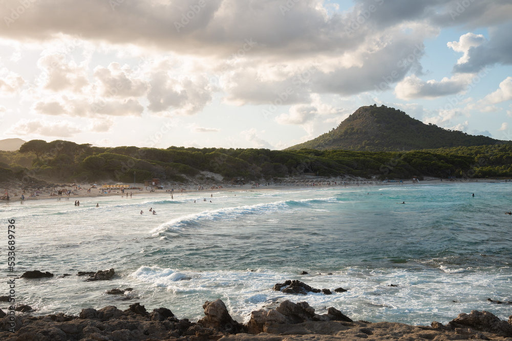 The waves splash on beach of Cala Agulla (Cala Ratjada) on Mallorca in the mediterranean sea. Blue water and a green mountain in the background. People bathing, surfers surfing. Tropical paradise.