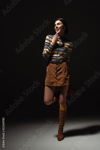 full length of excited young woman in turtleneck with skirt and footwear posing on black.