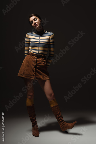 full length of young woman in turtleneck with skirt and autumnal boots posing on black.