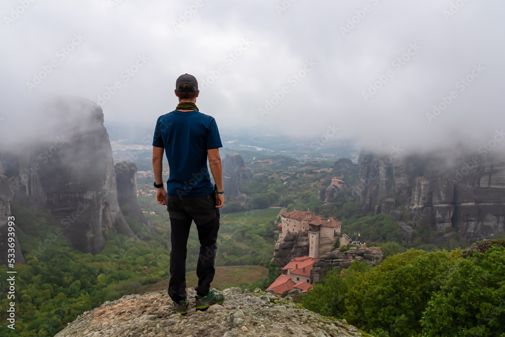 Man with scenic view of Holy Monastery of Rousanos appearing from fog, Kalambaka, Meteora, Thessaly, Greece, Europe. Mist atmosphere in dramatic landscape. Landmark build on unique rock formations