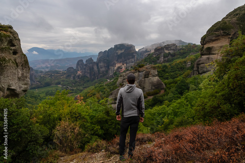 Man with scenic view of Holy Monastery of Rousanos and Holy Monastery St Nicholas Anapafsas appearing from fog, Kalambaka, Meteora, Thessaly, Greece, Europe. Misty atmosphere in dramatic landscape