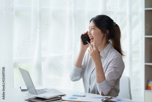 Charming and smiling Asian business woman Talk to customers on mobile phones and use their laptops to do office work.