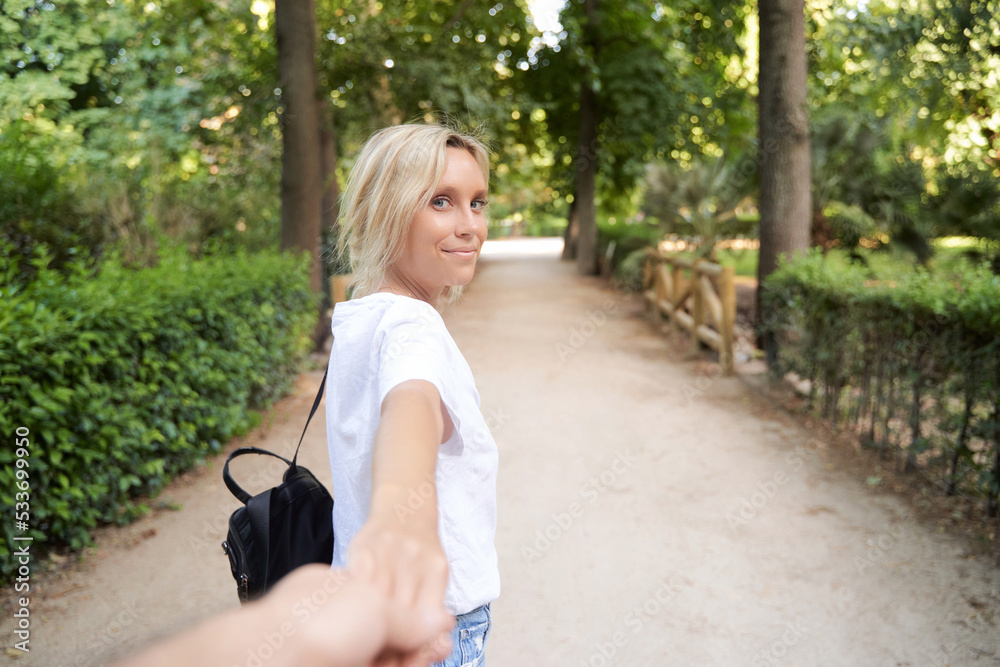 Young woman looking at camera and smiles while holding hand and leading someone on a walk outdoors in a park.