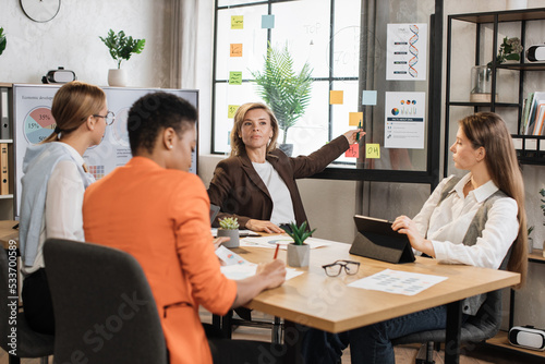 Senior blond financier woman pointing on board with paper sticks during speech on conference with multiracial female colleagues. Competent economists discussing company incomes at office room.