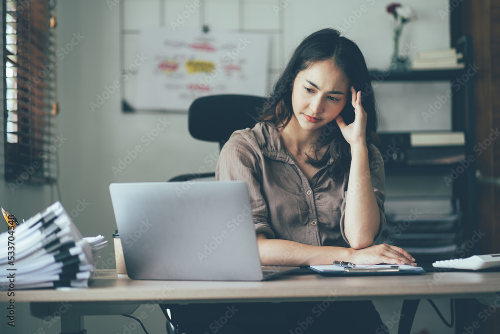 Portrait of a beautiful Asian woman looking at laptop screen while sitting at working desk in the office