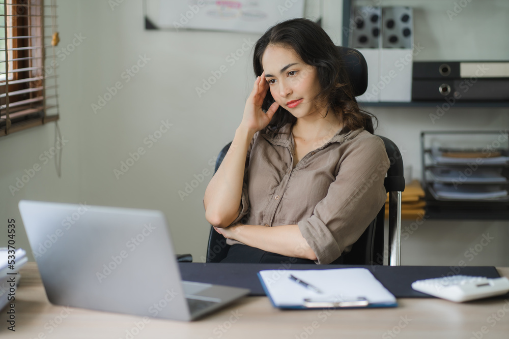 Portrait of a beautiful Asian woman looking at laptop screen while sitting at working desk in the office