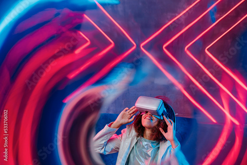 Smiling woman with VR glasses in front of neon light seen through glass photo