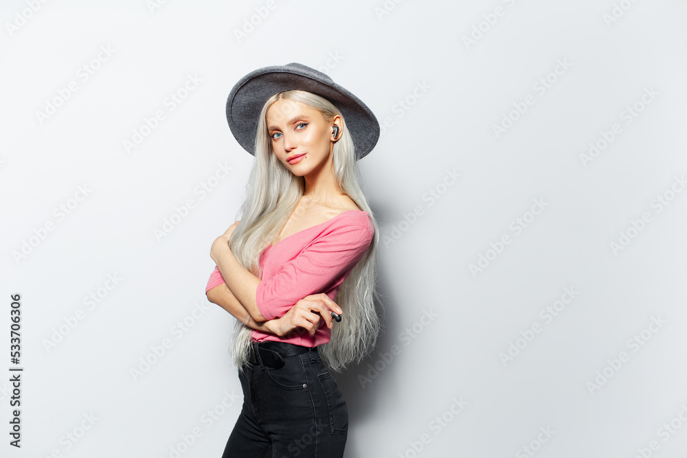 Studio portrait of young beautiful blonde girl with wireless earbuds in ear, wearing grey hat and pink shirt on white background. Copy space. Studio portrait of young beautiful blonde girl with wireless earbuds in ear, wearing grey hat and pink shirt on white background. Copy space.