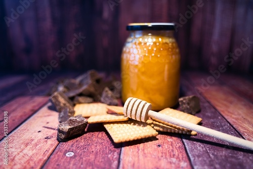 Jar of bee honey with a honey dipper, some cookies, and dark chocolate placed on a wooden surface
