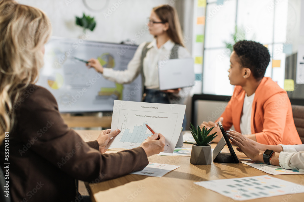 Fototapeta premium Focus on hands holding financial report. Female corporate director with laptop in hands having business meeting with international colleagues. Various diagrams, charts and technology at office.