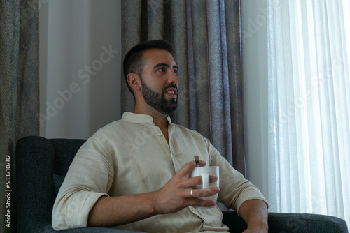 Young man in shirt holding a cup in the living room of his house happily