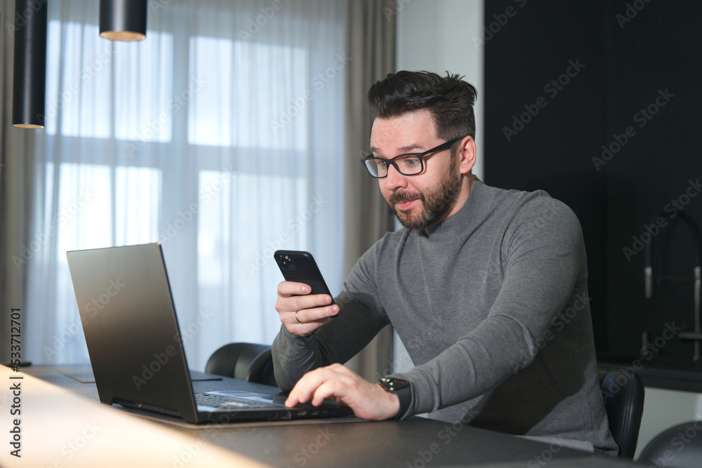 Portrait of surprised shocked young man with amazed face looking at screen of his smartphone in glasses, working on laptop computer at home 