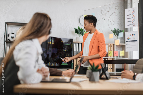 Multiracial female business people having briefing about financial situation at company. African woman pointing on monitor with various graphs and charts. Cooperation and brainstorming concept.