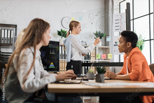 Caucasian blond woman in formal wear pointing on glass board with charts and graphs during working conference. Multi ethnic colleagues sitting at desk and listening female partner.