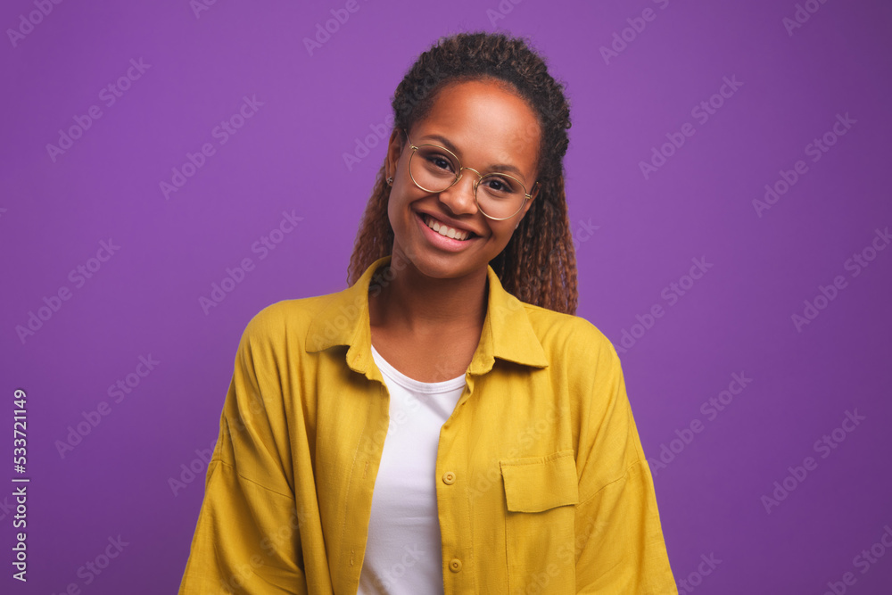 Young cheerful beautiful African American woman student wearing glasses smiling sincerely tilt head to side and look at you dressed in casual style stands on purple plain background Young cheerful beautiful African American woman student wearing glasses smiling sincerely tilt head to side and look at you dressed in casual style stands on purple plain background
