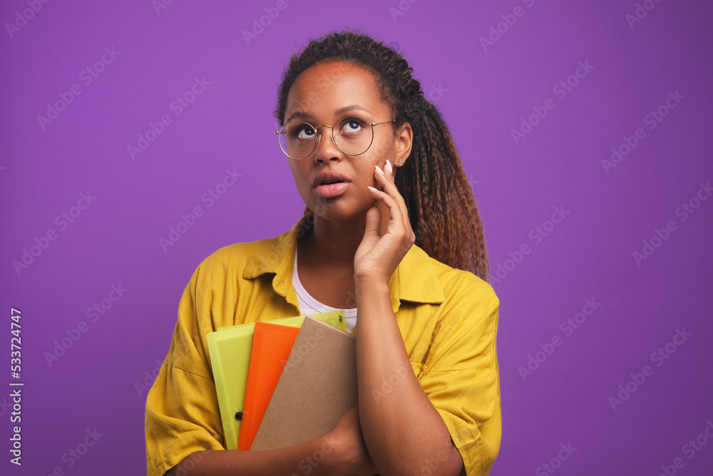 Young perplexed casual African American woman freelancer holds folders with working documents in front of chest and looks up trying to remember outstanding tasks standing in purple studio. Ethnic girl Young perplexed casual African American woman freelancer holds folders with working documents in front of chest and looks up trying to remember outstanding tasks standing in purple studio. Ethnic girl