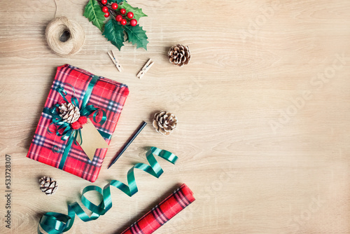 Flat lay of a Christmas gift box wrapped in scottish tartan pattern paper with wrapping materials over a wooden background. Christmas fever for presents preparation. Copy space.