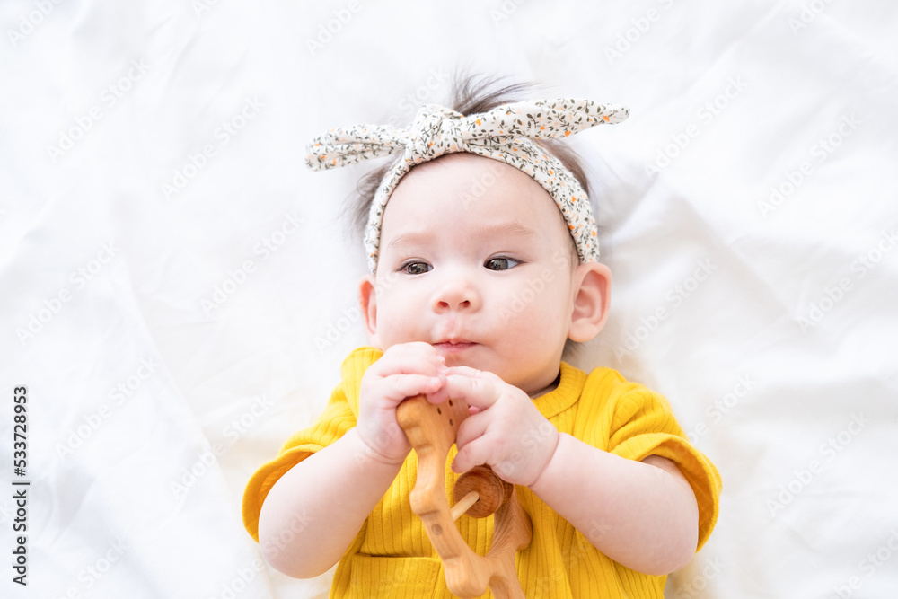 healthy asian baby girl in yellow bodysuit plays with wooden toy teether for teeth on white bedding healthy asian baby girl in yellow bodysuit plays with wooden toy teether for teeth on white bedding